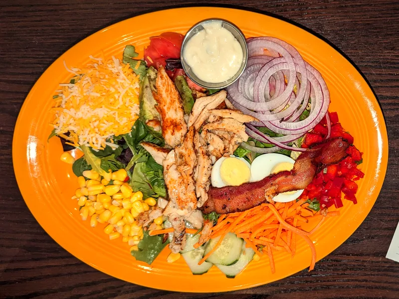 Cobb salad on an orange plate with rows of grilled chicken, chopped egg, bacon, cheese, corn, red onion, cucumber, avocado and tomato over greens