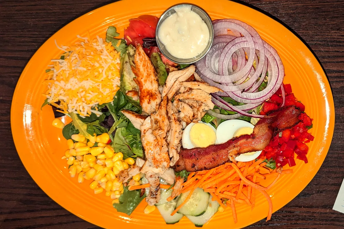 Cobb salad on an orange plate with rows of grilled chicken, chopped egg, bacon, cheese, corn, red onion, cucumber, avocado and tomato over greens