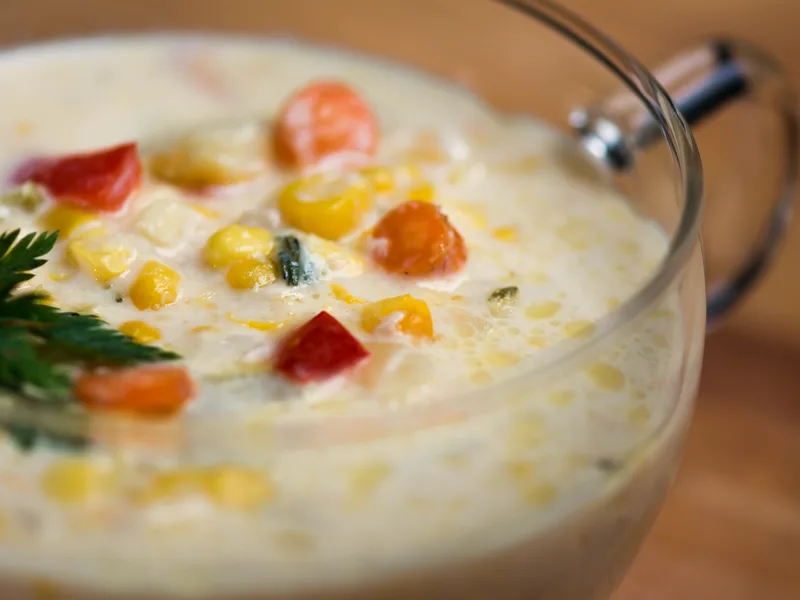 A close-up of creamy corn chowder in a clear glass mug, with corn kernels, diced red pepper and carrot and a parsley garnish