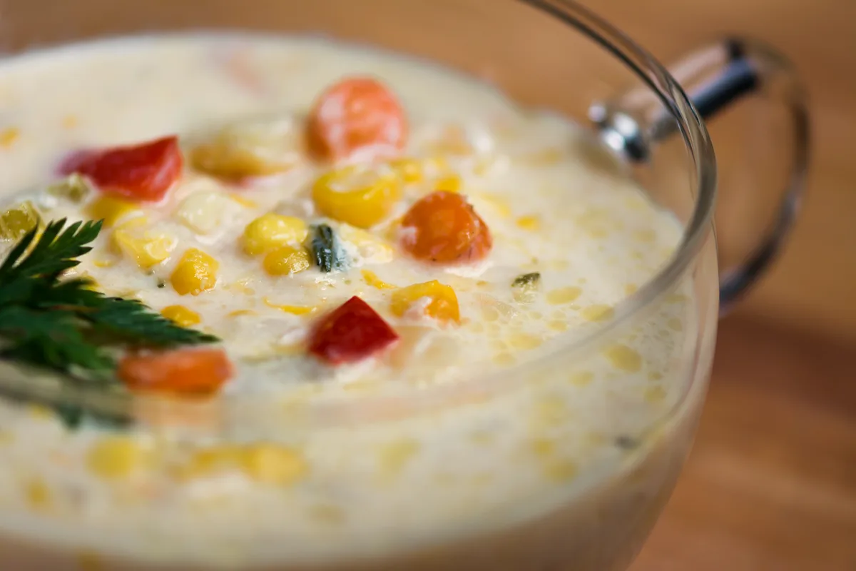 A close-up of creamy corn chowder in a clear glass mug, with corn kernels, diced red pepper and carrot and a parsley garnish