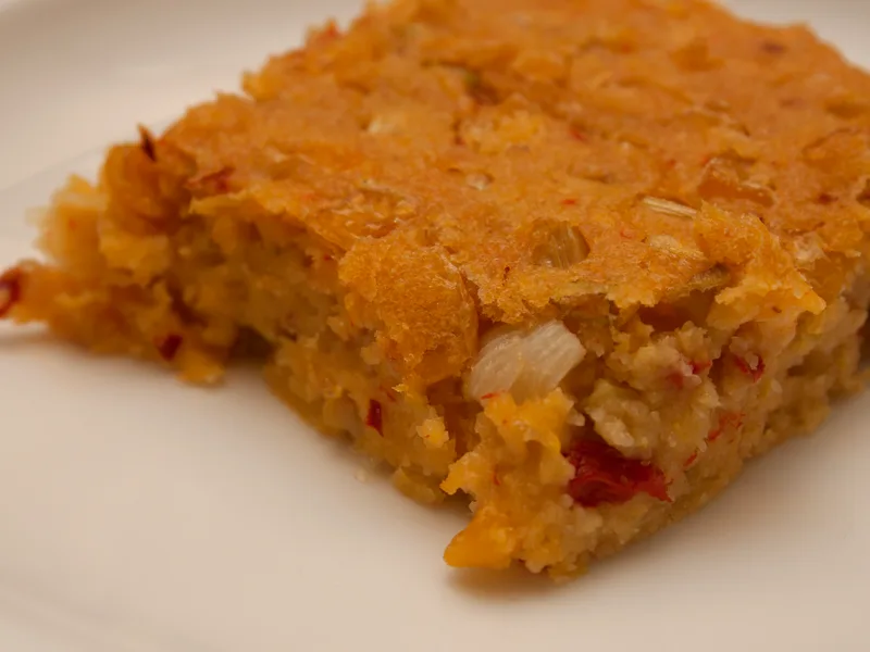 A square slice of golden Southwestern corn pudding on a white plate, with visible corn kernels, diced onion and red pepper pieces