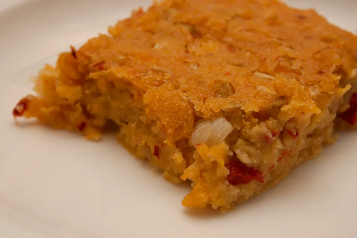 A square slice of golden Southwestern corn pudding on a white plate, with visible corn kernels, diced onion and red pepper pieces