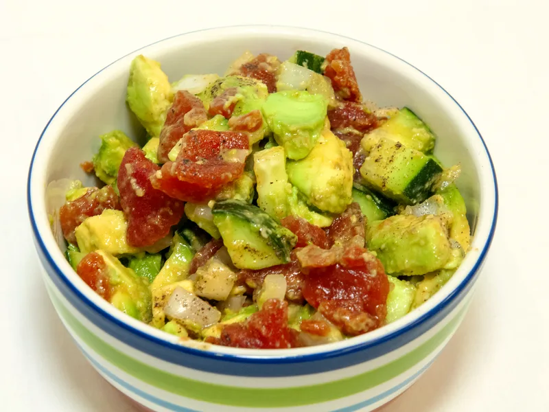 A bowl of diced cucumber, tomato and avocado salad with red onion and black pepper in a striped bowl