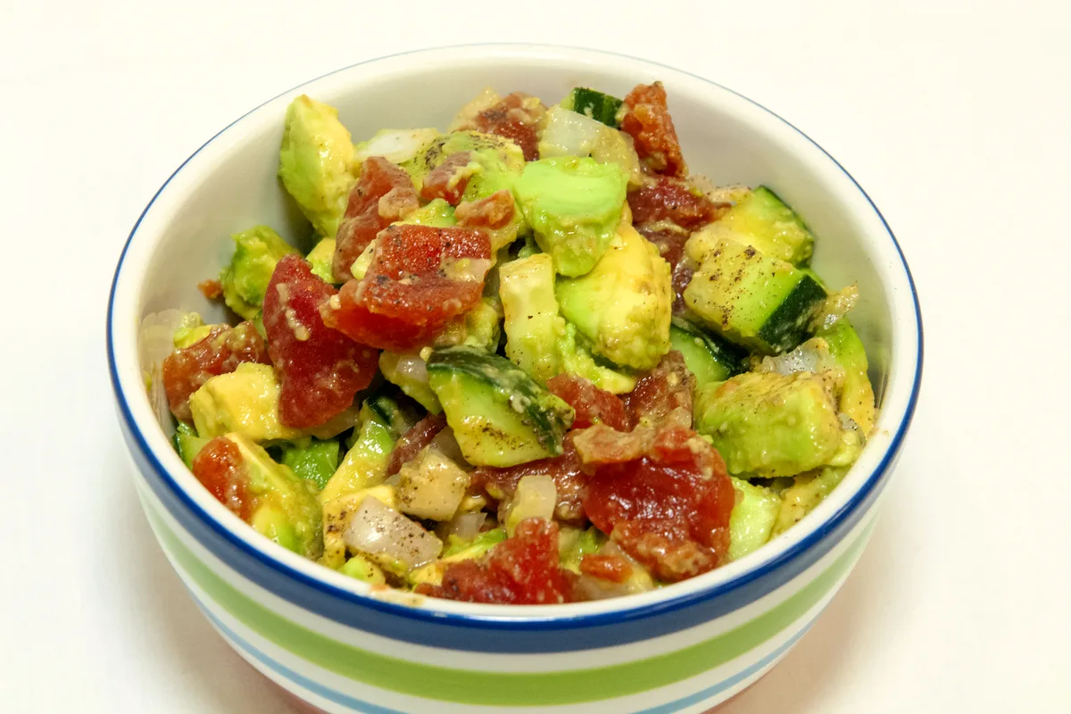 A bowl of diced cucumber, tomato and avocado salad with red onion and black pepper in a striped bowl