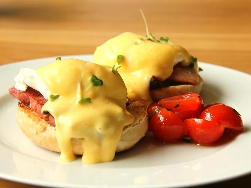 Two eggs Benedict on a white plate - toasted English muffin halves topped with ham, poached eggs and glossy yellow hollandaise sprinkled with chopped chives, with a small pile of roasted cherry tomatoes on the side