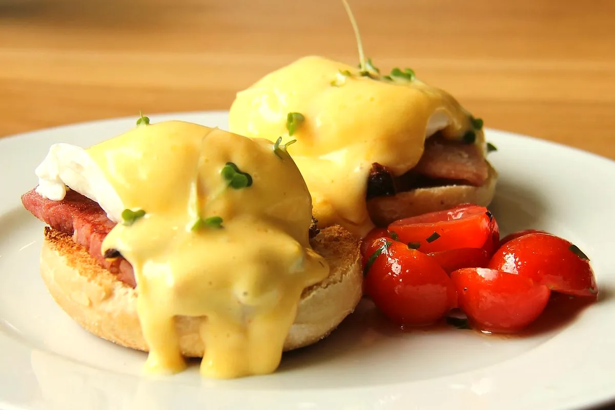 Two eggs Benedict on a white plate - toasted English muffin halves topped with ham, poached eggs and glossy yellow hollandaise sprinkled with chopped chives, with a small pile of roasted cherry tomatoes on the side