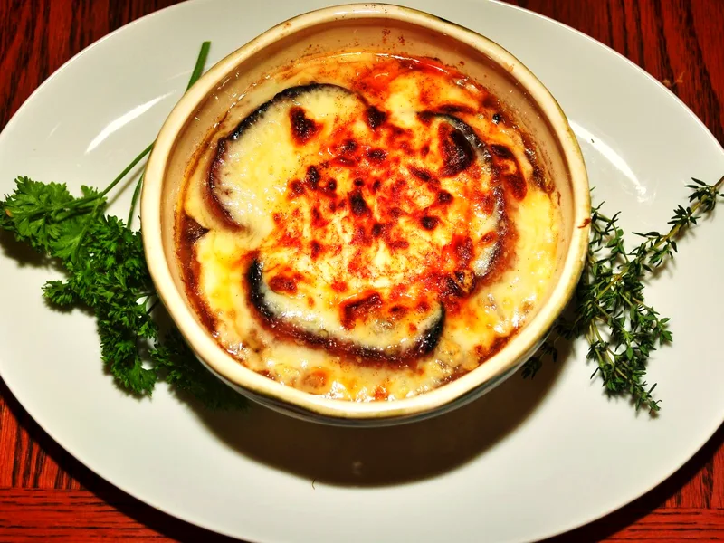 A white crock of slow cooker French onion soup with a bubbling, browned Gruyere crust over toasted baguette, garnished with parsley and thyme
