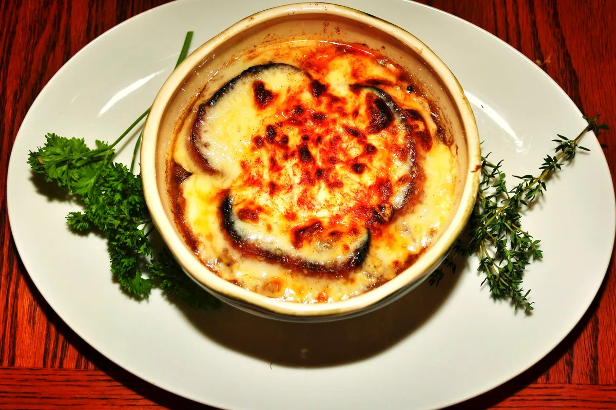 A white crock of slow cooker French onion soup with a bubbling, browned Gruyere crust over toasted baguette, garnished with parsley and thyme