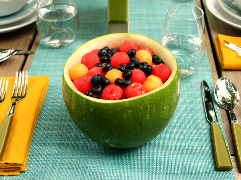 A hollowed melon half filled with red watermelon balls, orange cantaloupe balls and fresh blueberries, served on a blue placemat with table settings