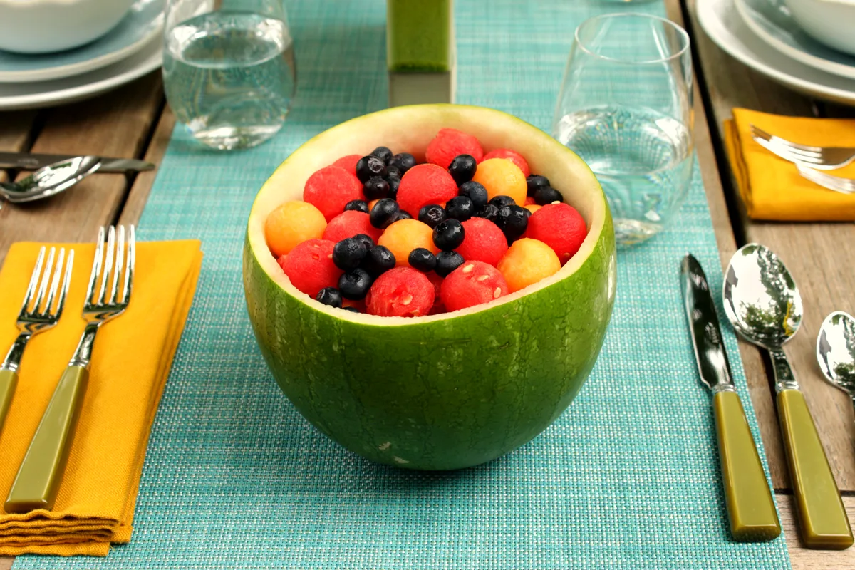 A hollowed melon half filled with red watermelon balls, orange cantaloupe balls and fresh blueberries, served on a blue placemat with table settings
