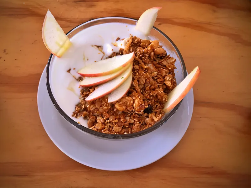 A glass bowl of white yogurt topped with a generous mound of golden granola and fanned fresh apple slices, with apple wedges at the rim, on a white saucer over a wooden table