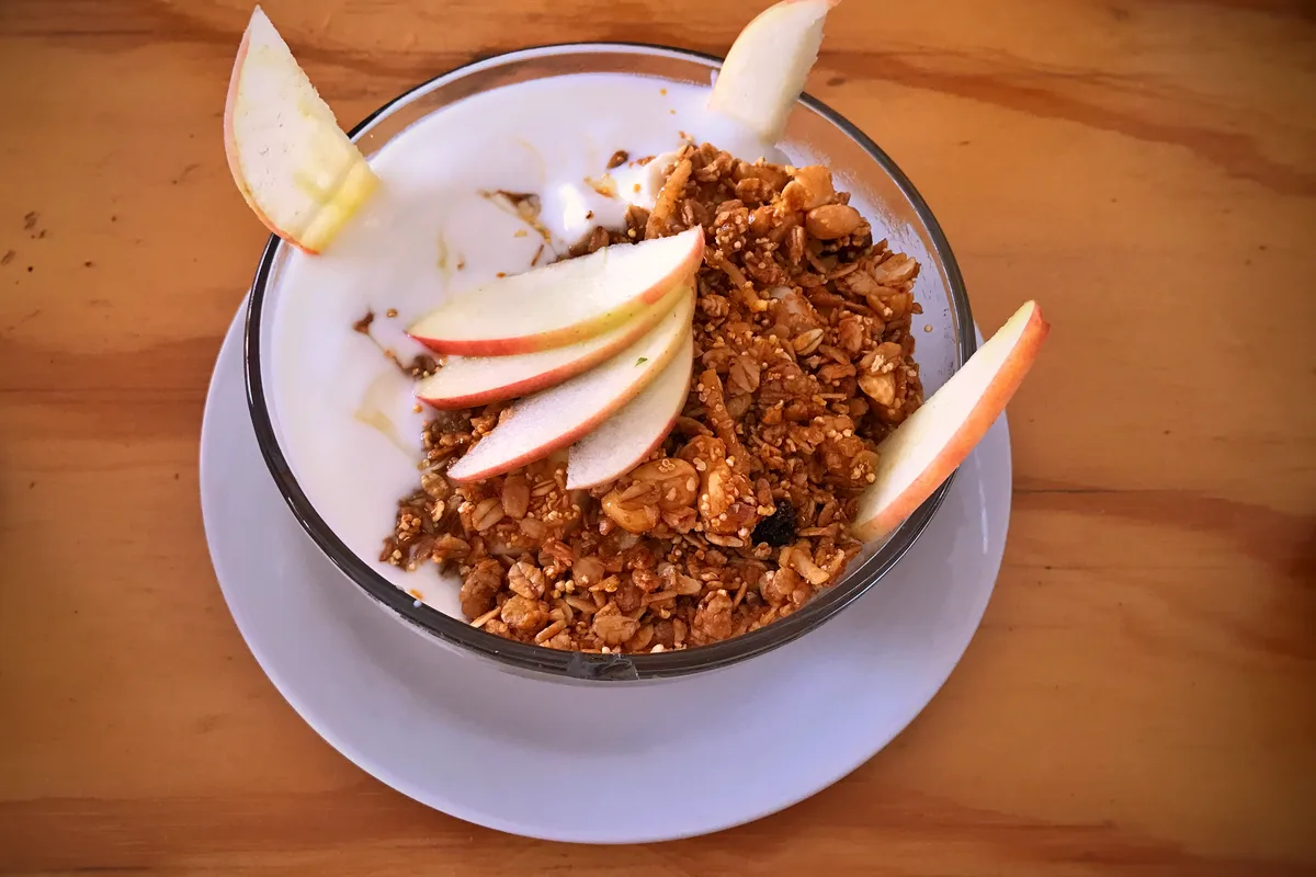 A glass bowl of white yogurt topped with a generous mound of golden granola and fanned fresh apple slices, with apple wedges at the rim, on a white saucer over a wooden table