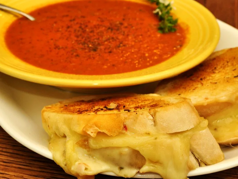 Two grilled cheese sandwich halves with melted cheese pulling between them on a white plate, in front of a yellow bowl of smooth roasted tomato soup sprinkled with black pepper and a thyme sprig