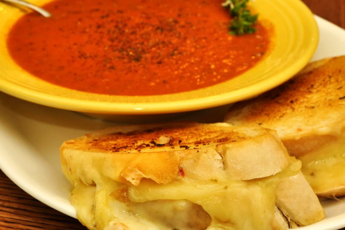 Two grilled cheese sandwich halves with melted cheese pulling between them on a white plate, in front of a yellow bowl of smooth roasted tomato soup sprinkled with black pepper and a thyme sprig