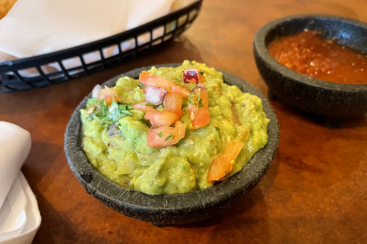 Easy homemade guacamole in a stone bowl topped with diced tomato, onion and cilantro, with salsa and tortilla chips beside it