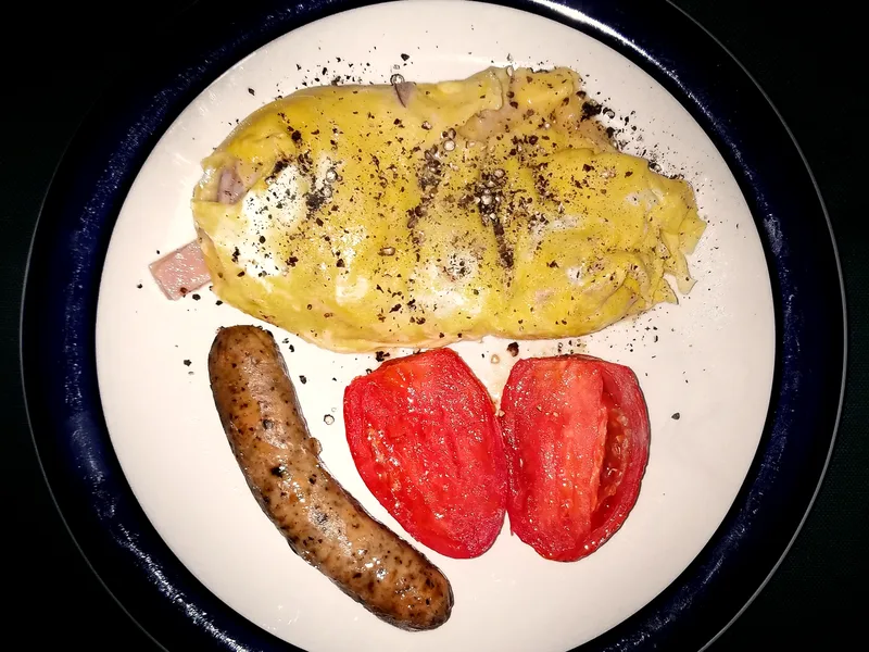 A folded ham and cheese omelette dusted with cracked black pepper on a white plate with a dark blue rim, served with one browned sausage link and two roasted plum tomato halves, seen from above
