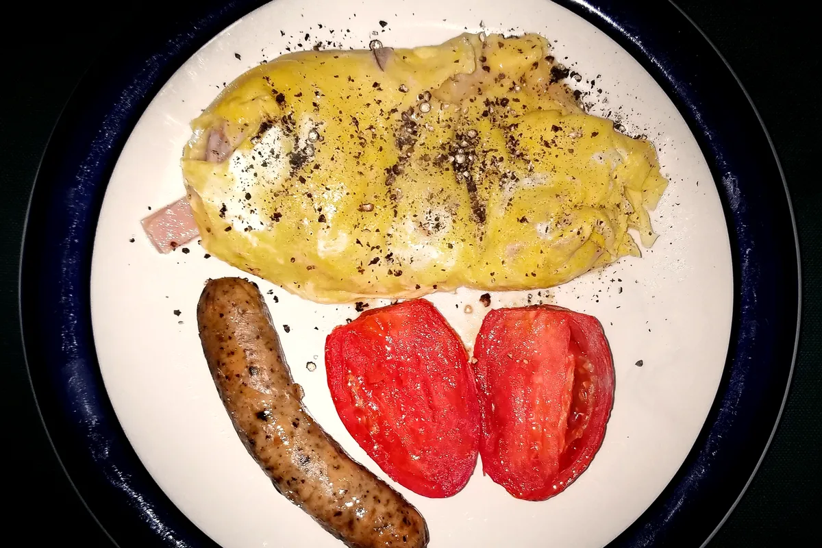 A folded ham and cheese omelette dusted with cracked black pepper on a white plate with a dark blue rim, served with one browned sausage link and two roasted plum tomato halves, seen from above