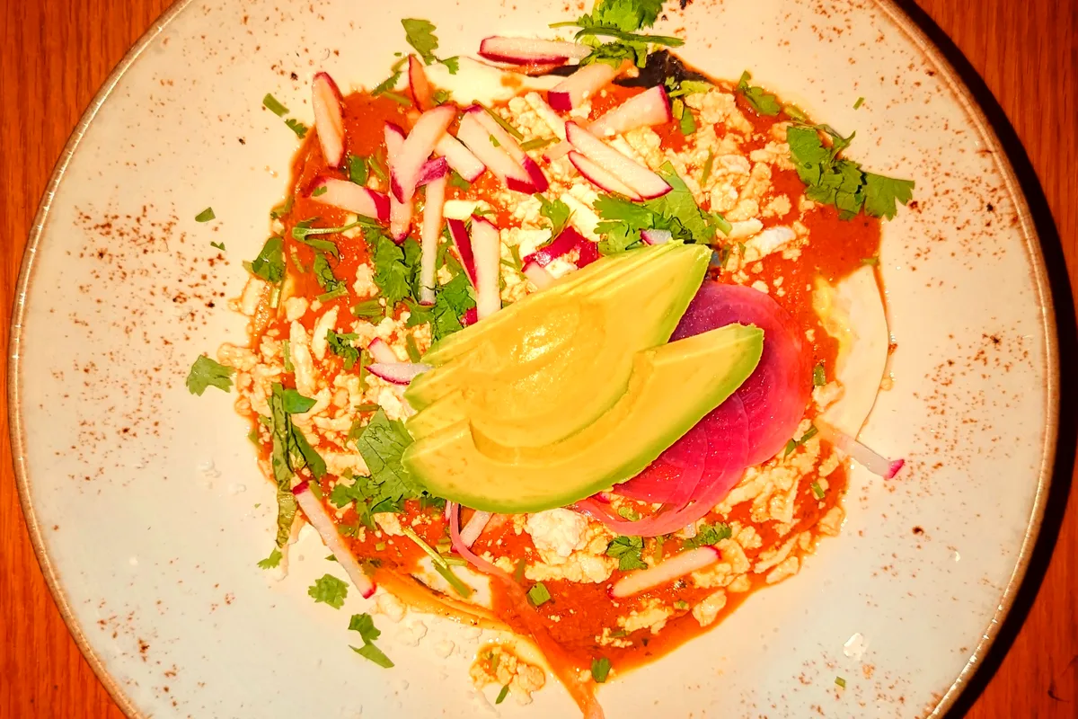 A plate of huevos rancheros with red ranchero sauce, crumbled cheese, sliced radish, cilantro, pickled red onion and avocado slices on a speckled plate