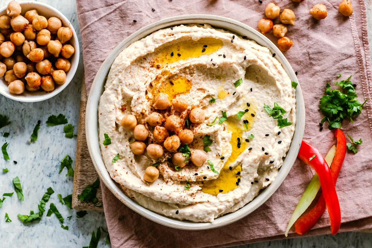 A bowl of smooth hummus swirled with pools of olive oil and topped with whole chickpeas, paprika and fresh herbs