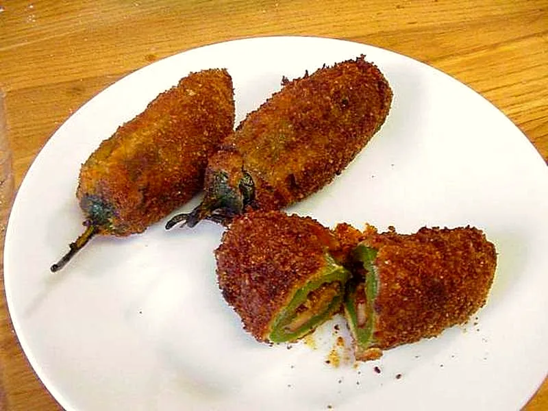 Three whole baked jalapeno poppers with a deep golden breadcrumb crust on a white plate, one cut open to show the green pepper wall and creamy filling inside