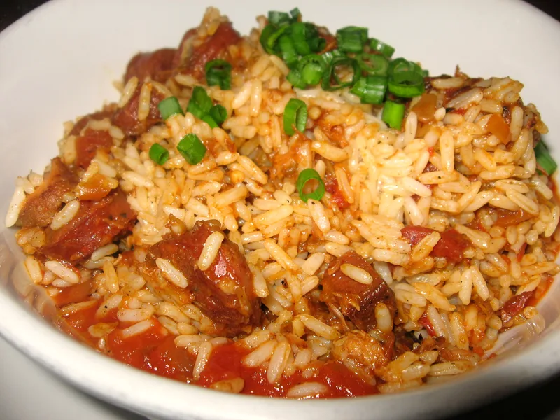 A white bowl of jambalaya with tomato-seasoned rice, chunks of smoked sausage and a scattering of sliced green onions on top
