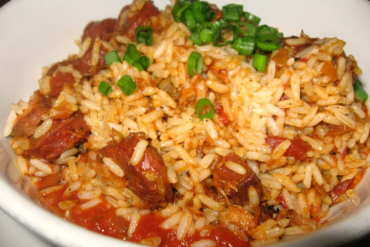 A white bowl of jambalaya with tomato-seasoned rice, chunks of smoked sausage and a scattering of sliced green onions on top