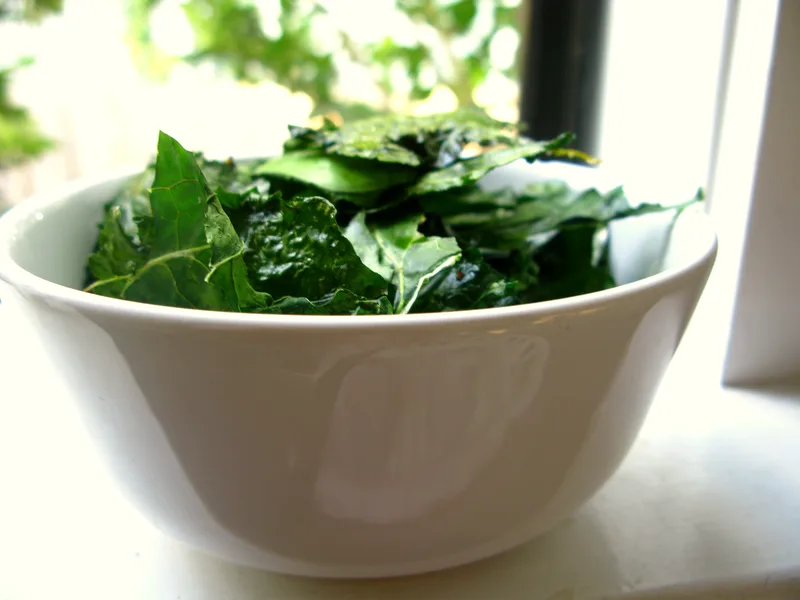 Crisp dark-green kale chips piled in a white bowl by a bright window