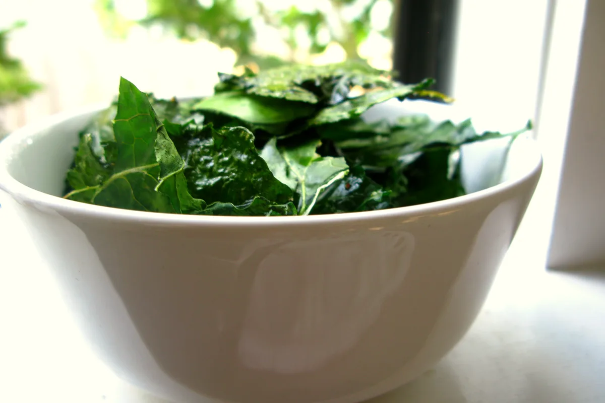Crisp dark-green kale chips piled in a white bowl by a bright window