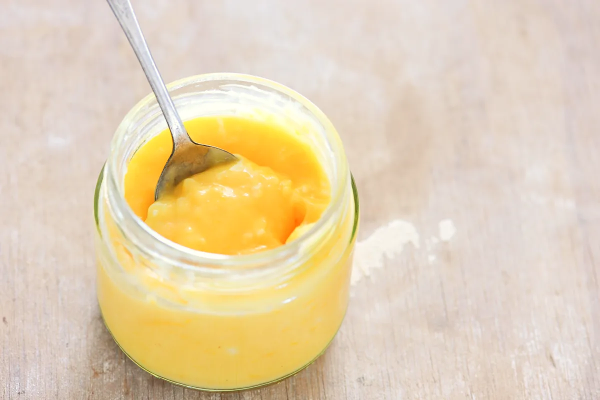 Open jar of homemade lemon curd with a spoon lifting a thick, glossy spoonful