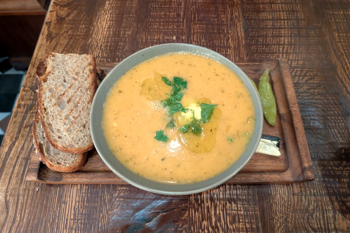 A bowl of smooth golden lentil soup topped with fresh herbs and olive oil, with slices of crusty bread alongside on a wooden board