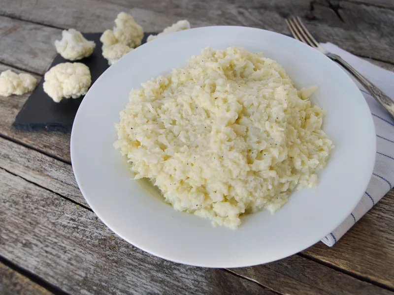 Plate of creamy garlic mashed cauliflower garnished with chives, with a whole cauliflower and florets beside it