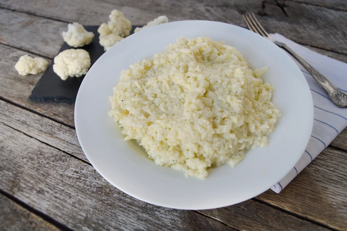 Plate of creamy garlic mashed cauliflower garnished with chives, with a whole cauliflower and florets beside it