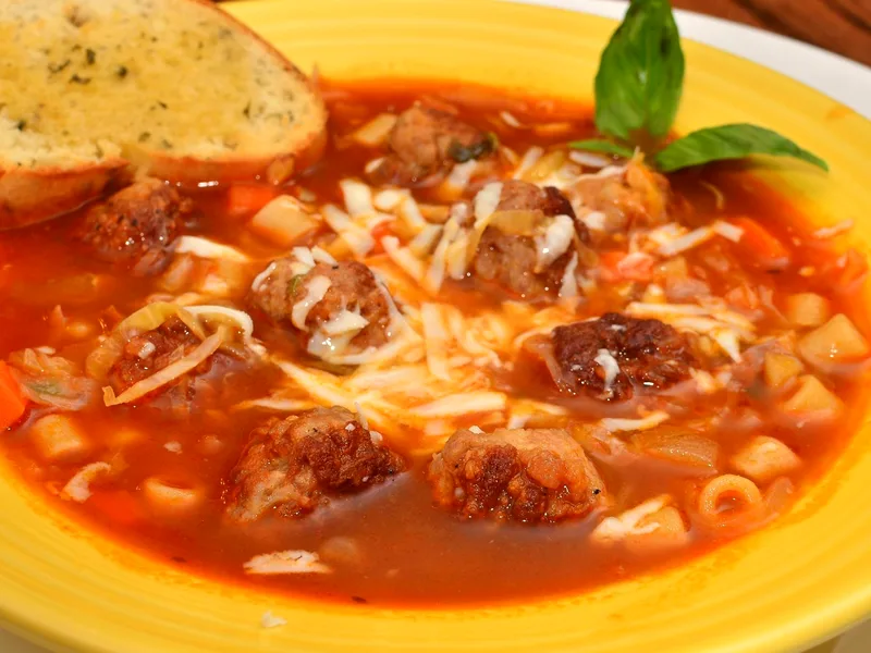 A yellow bowl of Italian meatball soup in a rich red tomato broth with browned meatballs, small pasta, diced carrots and potatoes and shredded melted cheese, garnished with basil leaves and a slice of garlic toast on the rim