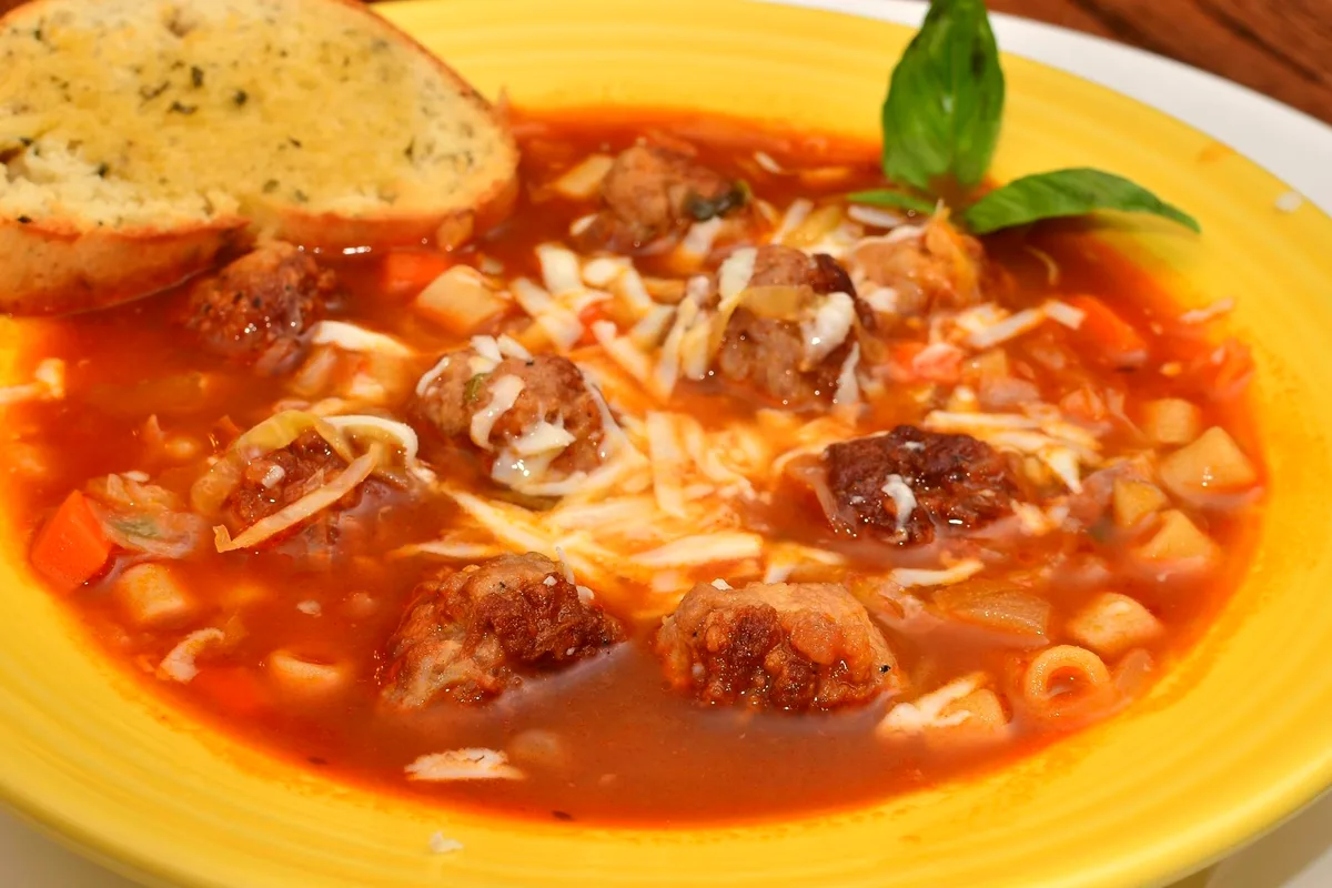 A yellow bowl of Italian meatball soup in a rich red tomato broth with browned meatballs, small pasta, diced carrots and potatoes and shredded melted cheese, garnished with basil leaves and a slice of garlic toast on the rim