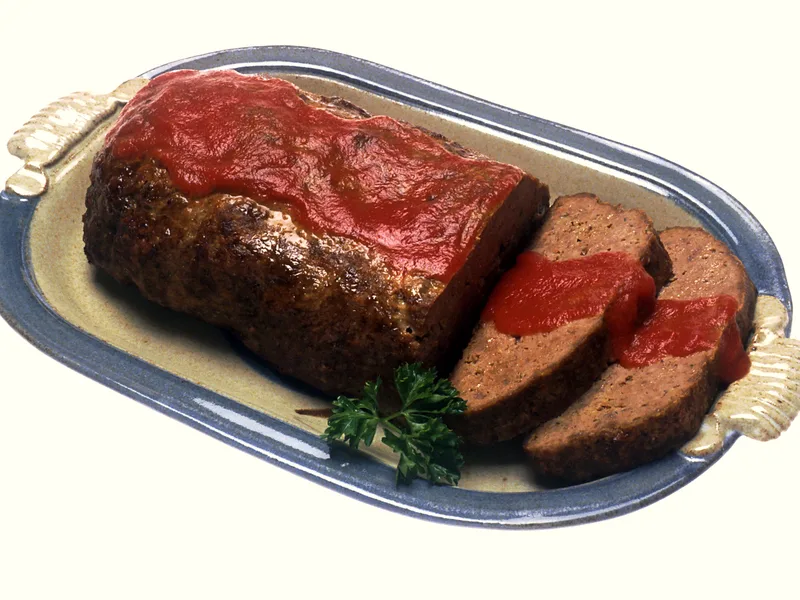 A classic meatloaf with a glossy red tomato glaze on an oval platter, two slices cut and a sprig of parsley alongside