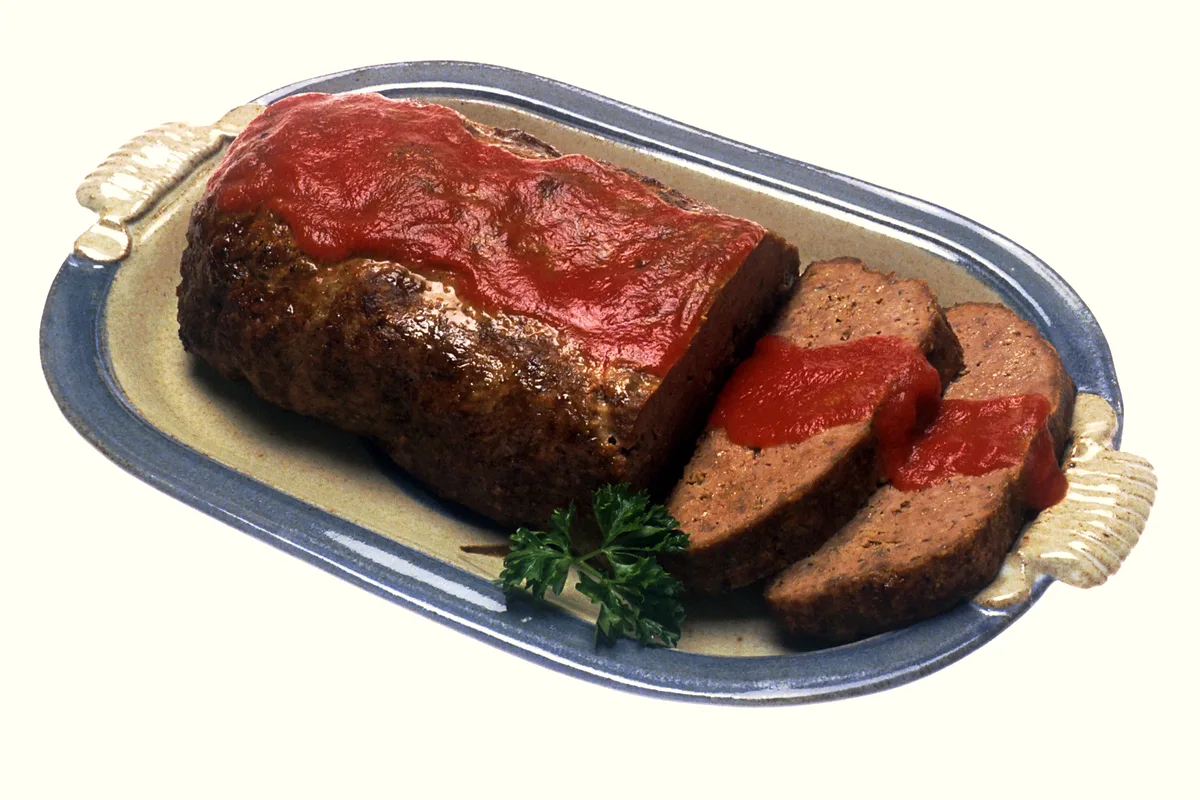 A classic meatloaf with a glossy red tomato glaze on an oval platter, two slices cut and a sprig of parsley alongside