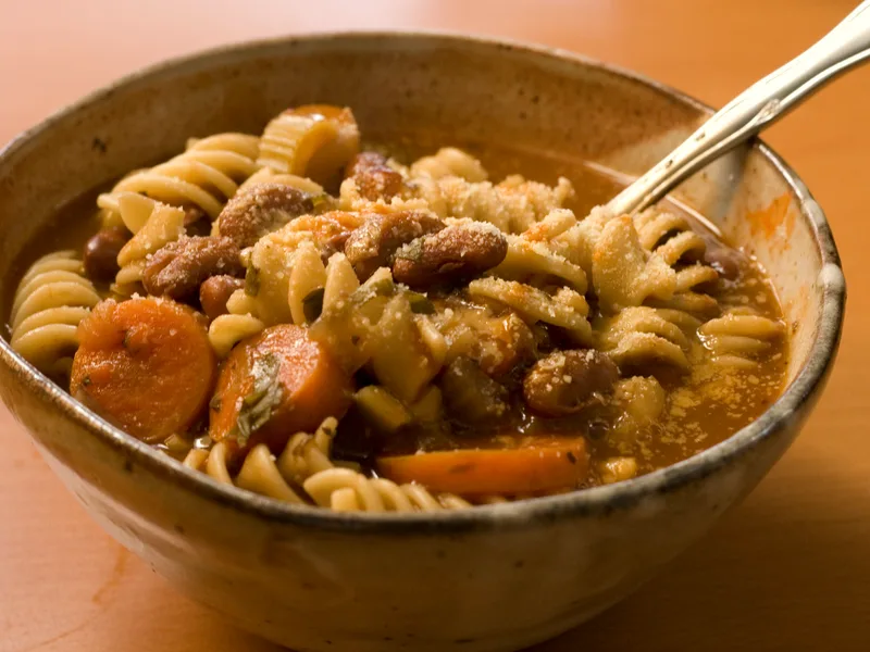 A rustic bowl of minestrone soup with rotini pasta, kidney beans, carrot coins and celery in tomato broth, dusted with Parmesan