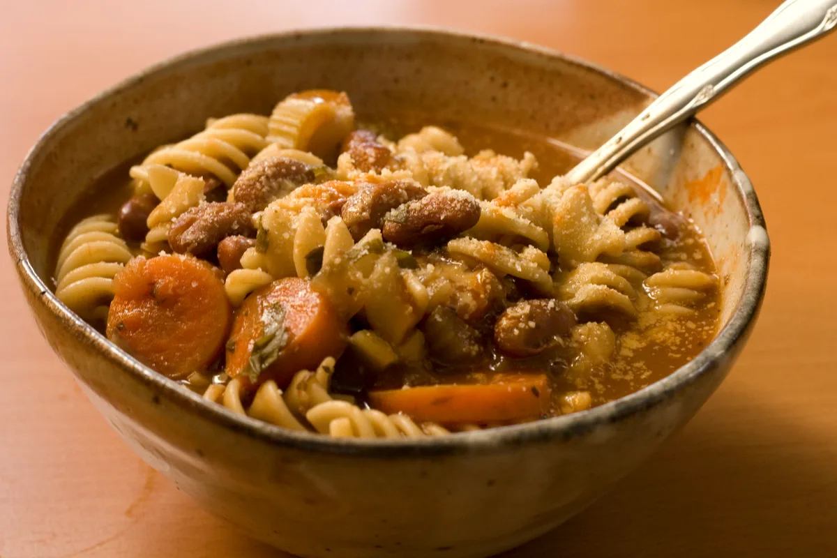 A rustic bowl of minestrone soup with rotini pasta, kidney beans, carrot coins and celery in tomato broth, dusted with Parmesan
