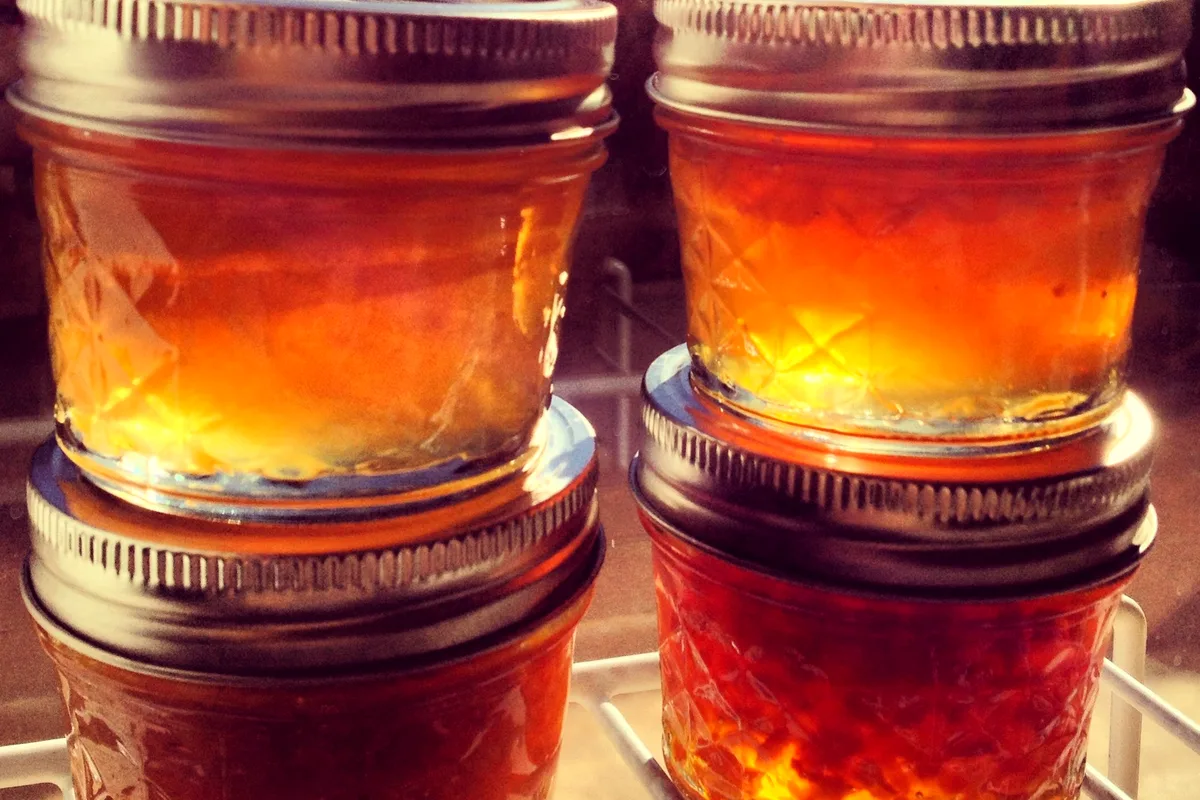 Four small sealed jars of golden-orange marmalade stacked on a rack, glowing amber in the light