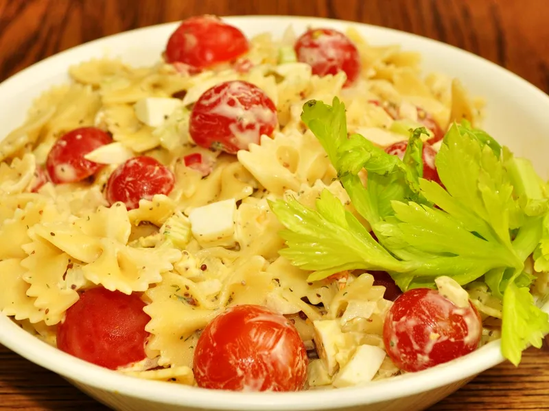 Bowl of creamy bowtie pasta salad with whole cherry tomatoes, chopped egg and celery, garnished with celery leaves