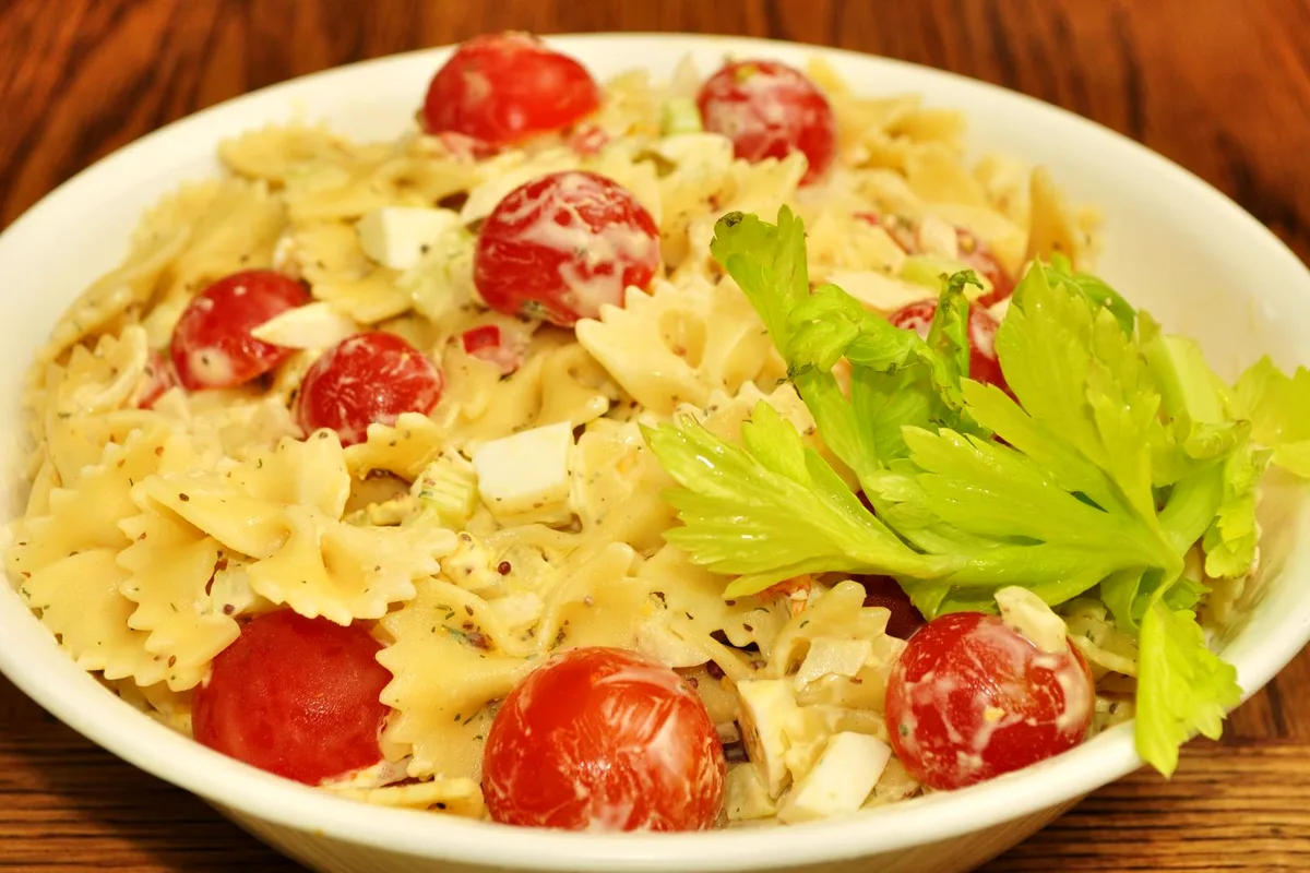 Bowl of creamy bowtie pasta salad with whole cherry tomatoes, chopped egg and celery, garnished with celery leaves