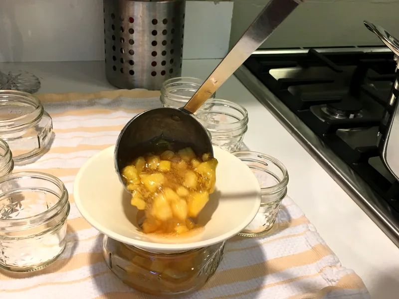 Ladling chunky golden peach jam through a canning funnel into small glass jars on a striped towel beside the stove