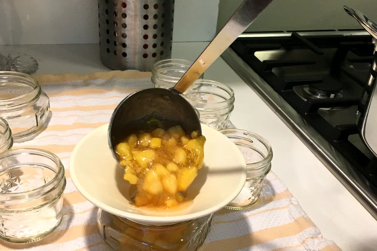 Ladling chunky golden peach jam through a canning funnel into small glass jars on a striped towel beside the stove