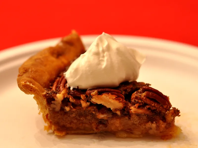 A slice of pecan pie on a white plate showing a gooey brown filling, a top layer of whole pecans and a flaky golden crust, topped with a large dollop of whipped cream
