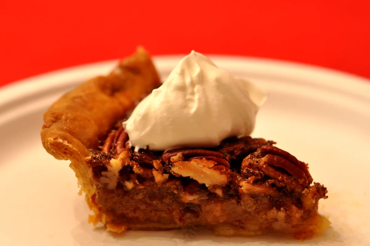A slice of pecan pie on a white plate showing a gooey brown filling, a top layer of whole pecans and a flaky golden crust, topped with a large dollop of whipped cream