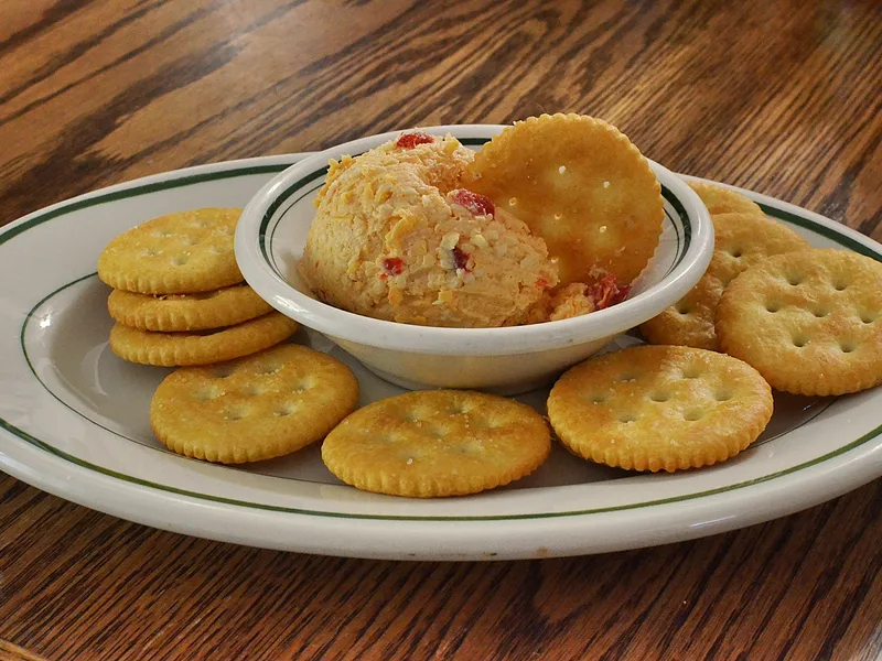 A small white bowl mounded with coarse orange pimento cheese spread studded with red pimento pieces, one round cracker dipped into it, ringed by round butter crackers on a white plate with a green rim