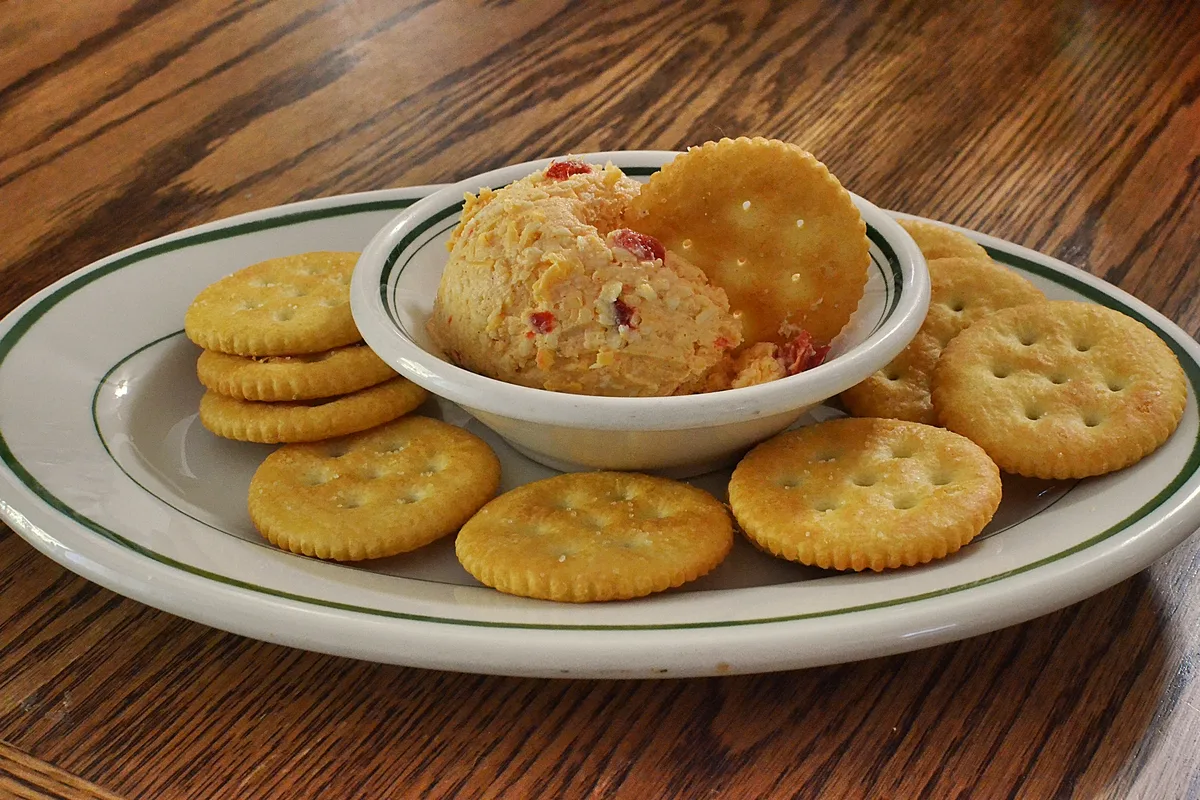A small white bowl mounded with coarse orange pimento cheese spread studded with red pimento pieces, one round cracker dipped into it, ringed by round butter crackers on a white plate with a green rim
