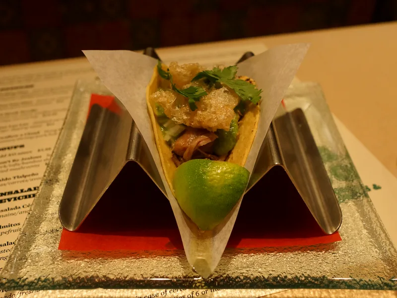 Carnitas taco in a holder topped with salsa verde, cilantro and crisp chicharron, with a lime wedge
