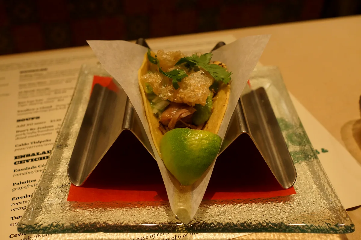 Carnitas taco in a holder topped with salsa verde, cilantro and crisp chicharron, with a lime wedge