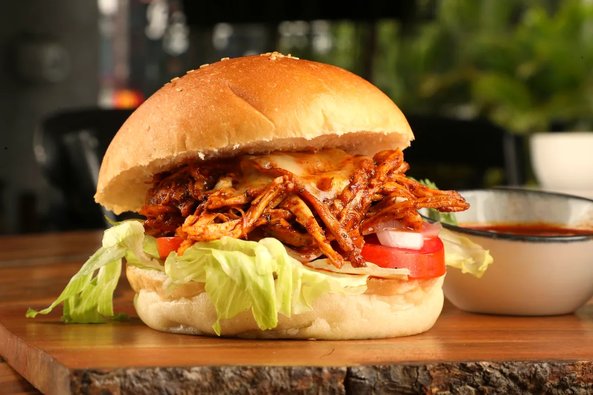 Slow cooker BBQ pulled chicken piled on a bun with coleslaw, with extra sauce in a small bowl
